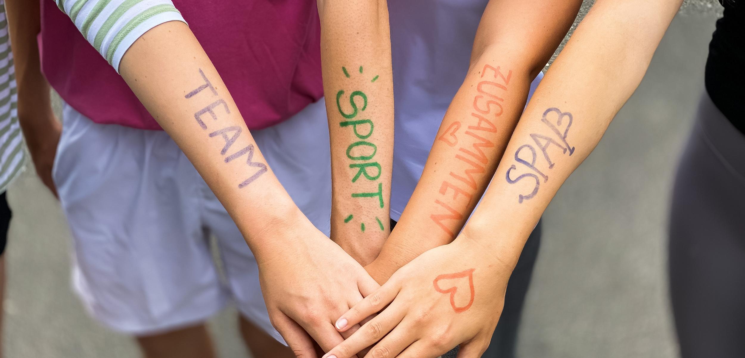 Vier Arme liegen übereinander, mit farbiger Schrift: „TEAM“, „SPORT“, „ZUSAMMEN“, „SPASS“ auf der Haut.