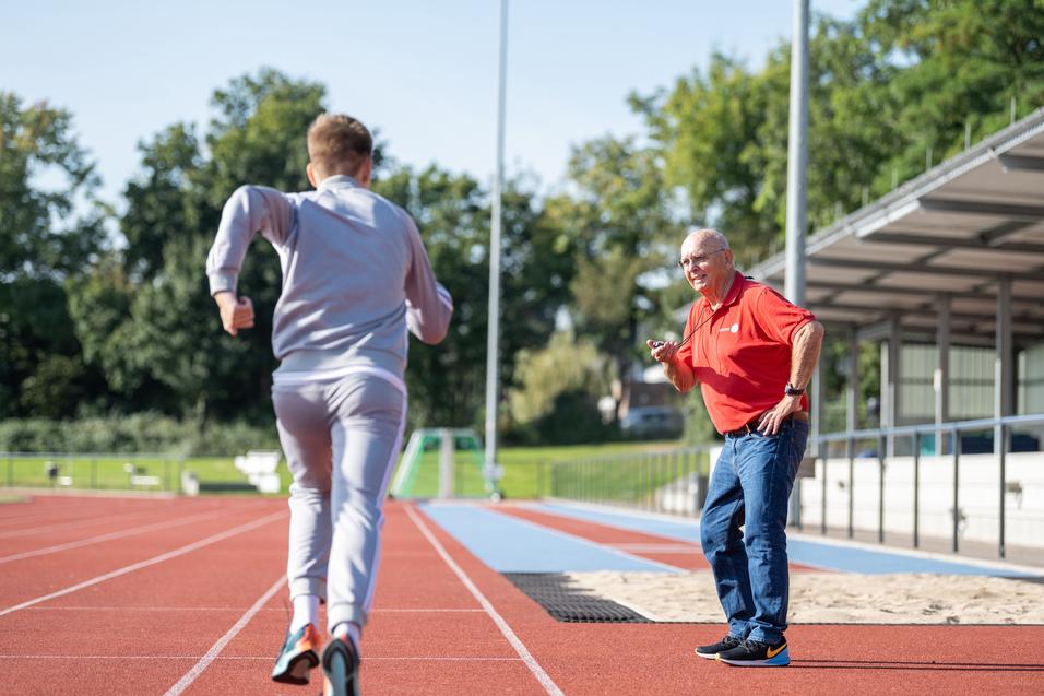 Trainer beobachtet einen Läufer auf der Bahn, während er eine Stoppuhr in der Hand hält. Der Sportler trägt sportliche Kleidung.